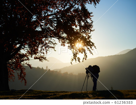 Hiker tourist man with camera on grassy valley on background of mountain landscape under big tree. 61236383