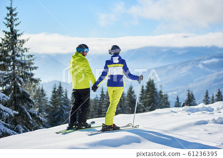 Back view of happy couple skiers standing on mountain edge, holding hands, enjoying winter mountain panoramic landscape 61236395