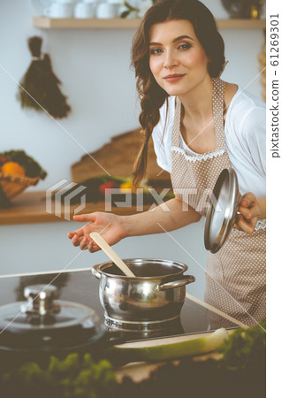 Young brunette woman cooking soup in kitchen. Housewife holding wooden spoon in her hand. Food and health concept Young brunette woman cooking soup in kitchen. Housewife holding wooden spoon in her hand. Food and health concept 61269301