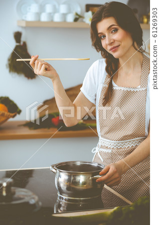 Young brunette woman cooking soup in kitchen. Housewife holding wooden spoon in her hand. Food and health concept Young brunette woman cooking soup in kitchen. Housewife holding wooden spoon in her hand. Food and health concept 61269303
