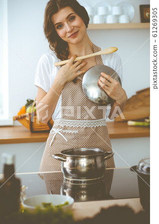 Young brunette woman cooking soup in kitchen. Housewife holding wooden spoon in her hand. Food and health concept Young brunette woman cooking soup in kitchen. Housewife holding wooden spoon in her hand. Food and health concept 61269305