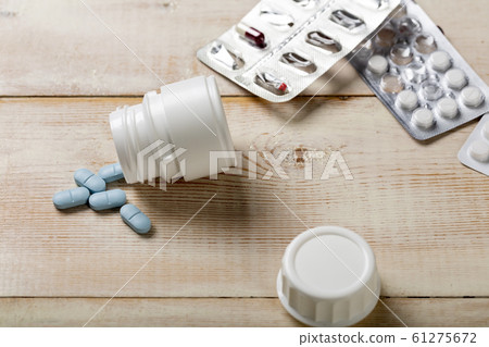 Blue pills spilled from a white plastic bottle. Different drug boxes and bottles in the background. Blue pills spilled from a white plastic bottle. Different drug boxes and bottles in the background. 61275672