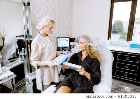 Professional cosmetology. Young woman cosmetologist standing near her female client while talking about beauty procedures and showing cosmetology medicines Professional cosmetology. Young woman cosmetologist standing near her female client while talking about beauty procedures and showing cosmetology medicines 61303015