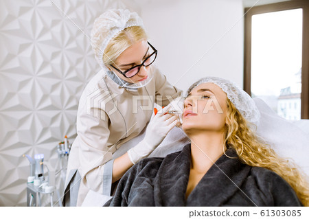 Attractive young blond woman is getting a rejuvenating facial injections at modern cosmetology clinic. Young female expert beautician is filling female wrinkles by hyaluronic acid Attractive young blond woman is getting a rejuvenating facial injections at modern cosmetology clinic. Young female expert beautician is filling female wrinkles by hyaluronic acid 61303085