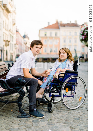 Young man and woman in wheelchair have fun and smiling on the bench outside. Old city street, love and disability concept Young man and woman in wheelchair have fun and smiling on the bench outside. Old city street, love and disability concept 61303141