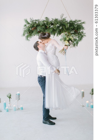 The groom lifted the bride up in his arms. The bride in luxury white wedding dress in the arms of the groom laughing. Wedding ceremony in light white room decorated with pine, flowers and candles. The groom lifted the bride up in his arms. The bride in luxury white wedding dress in the arms of the groom laughing. Wedding ceremony in light white room decorated with pine, flowers and candles. 61303379