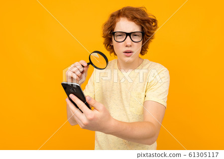 a teenager in glasses with a magnifier in his hand looks at a smartphone in his hand on a yellow a teenager in glasses with a magnifier in his hand looks at a smartphone in his hand on a yellow 61305117