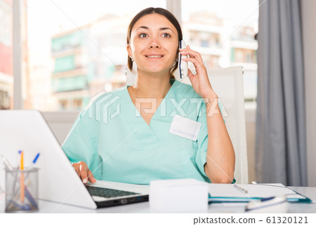 Practicioner woman in medical uniform talking by phone at laptop 61320121