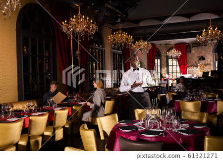 Polite waiter holding tray in restaurant with customers behind him 61321271
