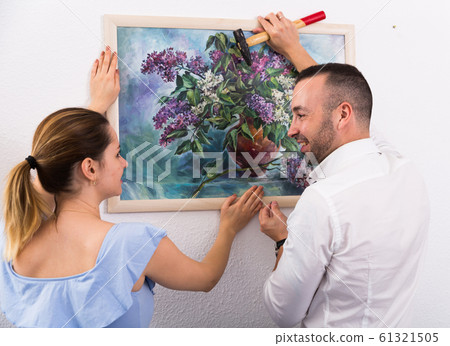 Man is hanging the picture on the wall together with his wife 61321505
