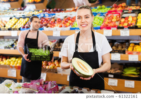 Young female seller with half of melon and man on background having box of green beans on the supermarket Young female seller with half of melon and man on background having box of green beans on the supermarket 61322161