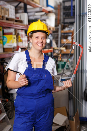Woman in uniform and helmet standing near racks in build store holding saw 61323239