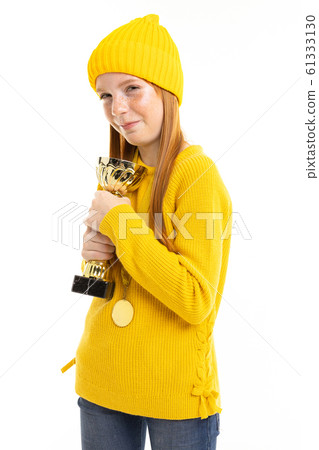 Happy teenager girl with red hair, yellow hoody and trousers holds a gold cup isolated on white 61333130