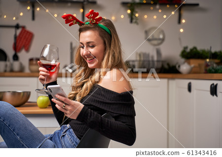 Happy woman with wine glass and phone in hand sitting in kitchen on new year Happy woman with wine glass and phone in hand sitting in kitchen on new year 61343416