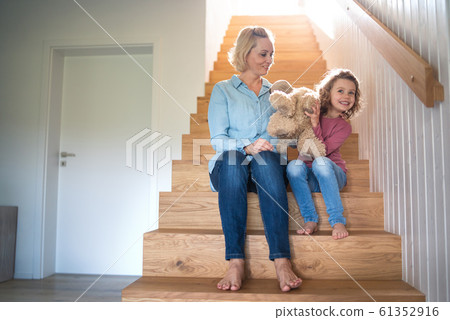 A cute small girl with mother indoors at home, sitting on staircase. 61352916