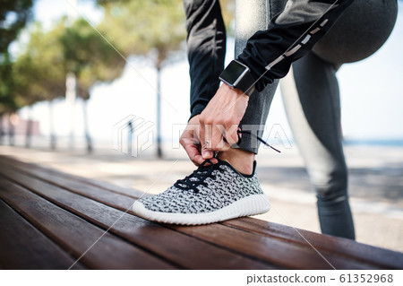 A young sportswoman with smartwatch outdoors in city, tying shoelaces. 61352968