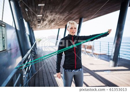 A young sportswoman with elastic band doing exercise outdoors in city. 61353013