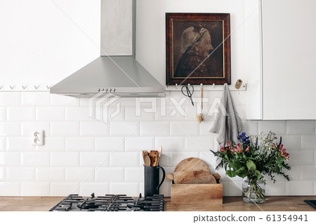 Modern eclectic kitchen interior. White brick wall with metro tiles, peg rails and oil painting. Wooden countertop, stainless steel hood and gas stove. Scandinavian design. Home staging, concept. Modern eclectic kitchen interior. White brick wall with metro tiles, peg rails and oil painting. Wooden countertop, stainless steel hood and gas stove. Scandinavian design. Home staging, concept. 61354941