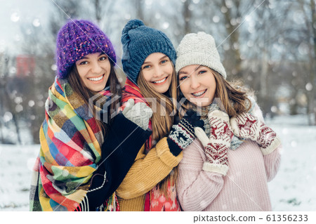 Three women friends outdoors in knitted hats having fun on a snowy cold weather. Group of young female friends outdoors in winter park. 61356233
