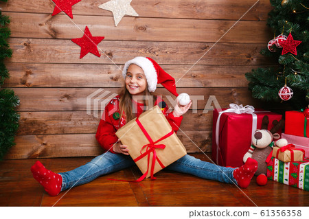 Cute little girl in Santa hat with present gift boxes sitting near tree at home. 61356358
