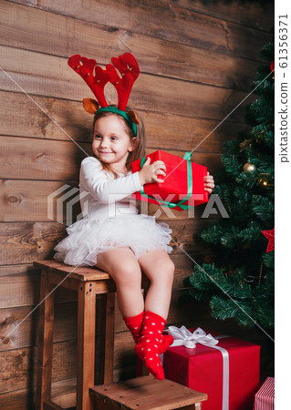 Smiling funny child in red deer horns holding Christmas gift in hand near Christmas tree at home Smiling funny child in red deer horns holding Christmas gift in hand near Christmas tree at home 61356371