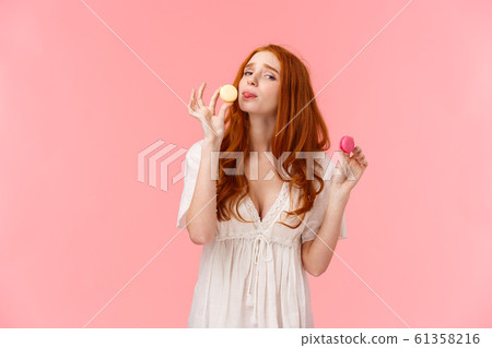 Girl on diet cant resist temptation eating sweets. Alluring funny and cute redhead female holding two tasty macarons want lick dessert, frowning and looking guilty camera, standing pink background 61358216