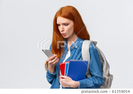 What is that. Confused and displesaed. intense troubled redhead teenage, college girl with backpack and notebooks walking university and reading strange text on smartphone, white background What is that. Confused and displesaed. intense troubled redhead teenage, college girl with backpack and notebooks walking university and reading strange text on smartphone, white background 61359054