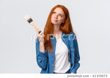Dreamy and indecisive cute redhead female in denim shirt, smirk and squinting thoughtful looking up imaging something, picturing ideas, holding paintbrush, thinking what color pick to paint 61359073