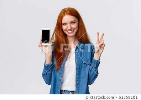 Cute and cheerful alluring redhead female in denim shirt, showing peace gesture smiling and holding smartphone, show app, company website or shopping internet store on mobile screen, white background Cute and cheerful alluring redhead female in denim shirt, showing peace gesture smiling and holding smartphone, show app, company website or shopping internet store on mobile screen, white background 61359101