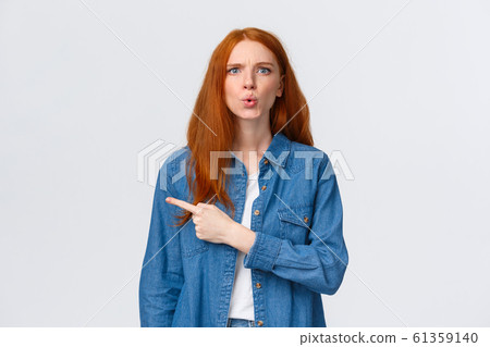 What is that. Alluring redhead girl in denim shirt, staring questioned, asking question and pointing finger left, frowning perplexed, dont know something, curious about product, white background 61359140