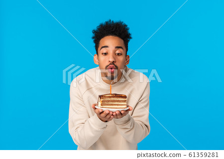 May all wishes fulfill. Cute stylish african-american african american guy with afro haircut, beard, holding birthday cake, blowing candle with folded lips as making wish, standing blue background 61359281