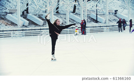 A young blonde woman skating on ice rink around other people outdoors - one leg up A young blonde woman skating on ice rink around other people outdoors - one leg up 61366160