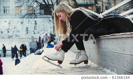 A young woman with blond hair tie up her figure skates before walking out on the rink A young woman with blond hair tie up her figure skates before walking out on the rink 61366184