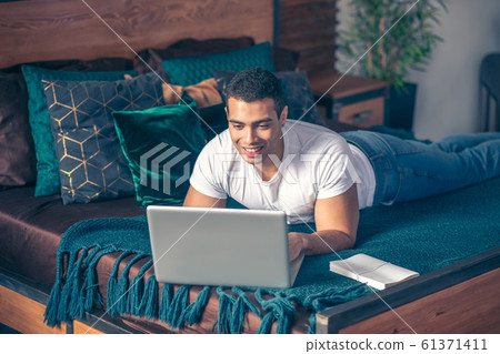 Smiling guy lying on the bed with a laptop. Smiling guy lying on the bed with a laptop. 61371411