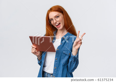 Sassy and cute, carefree happy redhead woman in glasses, denim shirt, showing peace sign and smiling camera cheerful as holding digital tablet, watching vlog online, working from distance 61375234