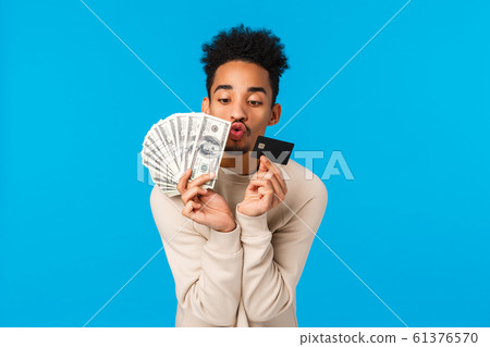 Waist-up portrait addicated and pleased african-american guy looking at precious money and credit card, like waisting cash on winter holidays sale season, kissing them, blue background Waist-up portrait addicated and pleased african-american guy looking at precious money and credit card, like waisting cash on winter holidays sale season, kissing them, blue background 61376570