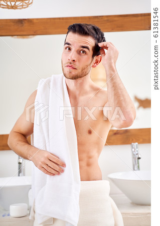 Young male with white towel on his shoulder standing by mirror in bath. Young male with white towel on his shoulder standing by mirror in bath. 61381546
