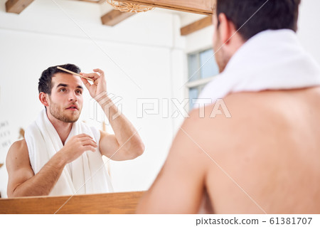 Unshaven man combing his hair while standing with white towel on neck near mirror in bathroom 61381707