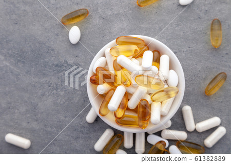 Assortment scattered pharmaceutical medicine vitamins, pills, drugs in bowl on gray background. White food dietary supplement hard shelled capsules fill with powder, salmon oil softgel Omega 3 61382889