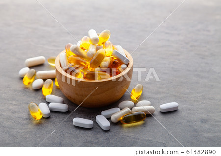 Assortment scattered pharmaceutical medicine tablets, pills, drugs in wooden bowl on gray background. White food dietary supplement hard shell capsules filled with powder, fish oil softgel Omega 3 61382890