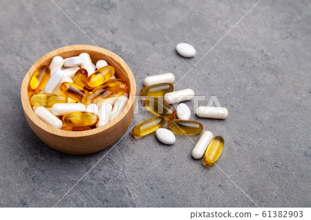 Assortment scattered pharmaceutical medicine tablets, pills, drugs in wooden bowl on gray background. White food dietary supplement hard shell capsules filled with powder, fish oil softgel Omega 3 61382903