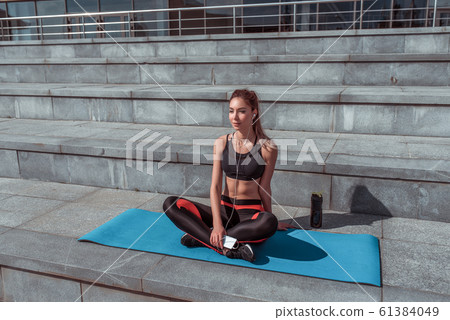 Beautiful girl resting after training on mat, summer city, sportswear, mobile phone. Enjoys music on headphones, listens audio recordings. Free space for copy text. Bottle shaker with protein water. 61384049