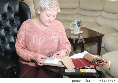 Portrait of an attractive mature short-haired woman in a pink sweater sits in a leather armchair in a furniture store and selects the color and upholstery material of the furniture. 61384757