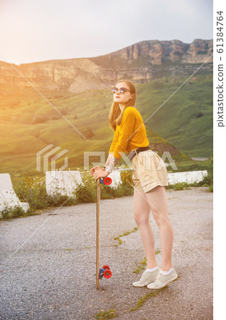 Beautiful and fashionable young woman in sunglasses and with a tattoo poses with a skateboard or longboard against the sunset sky 61384764