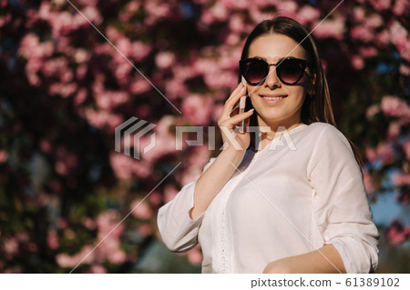 Portrait of attravtive young woman in white shirt and sunglasses use smartphone outside. Speel with someboby by the phone. Background of pink tree 61389102