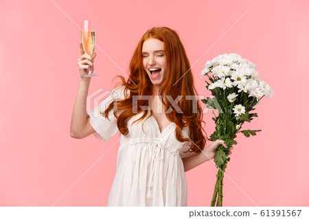 Excited and carefree good-looking redhead woman celebrating bachelorette party, scream yeah with closed eyes and relaxed expression, feeling awesome, holding bouquet flowers and raise champagne 61391567