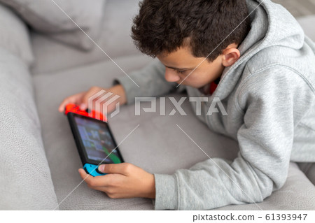 Teenager lies on a sofa and plays a game. He has a portable game console in his hands. How teens spend their vacations. Teenager lies on a sofa and plays a game. He has a portable game console in his hands. How teens spend their vacations. 61393947