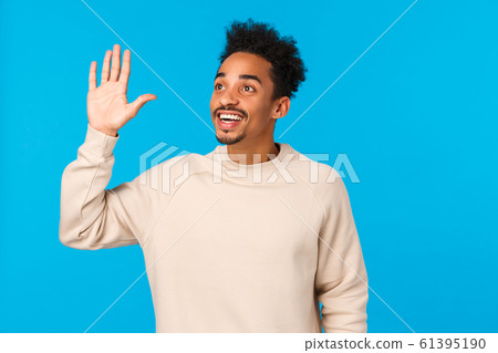 Hey hello, over here. Attractive friendly african-american hipster guy afro haircut, moustache, seeing friend in crowd, looking upper left corner, waving hand in hi gesture, smiling, blue background 61395190