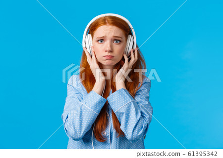 Girl about cry from emotional song. Upset redhead girlfriend suffering breakup, sobbing listening music in large white headphones, standing blue background in nightwear, unhappy 61395342