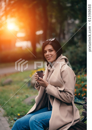 Beautiful young woman sitting on a bench drinking coffee enjoying in park 61398688